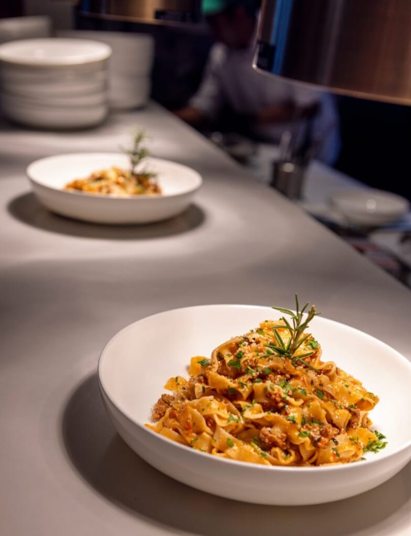 Two bowls of pasta with meat sauce and rosemary on a kitchen counter, with a chef in the background.