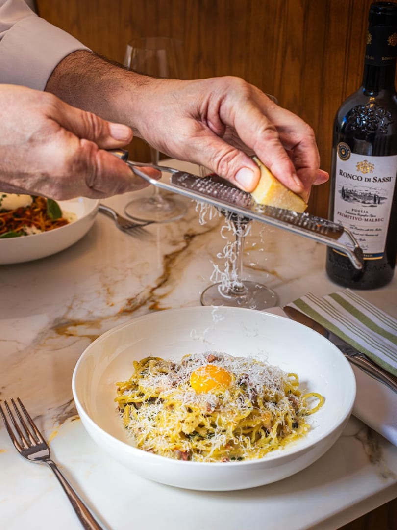 Hands grating cheese over a bowl of pasta carbonara with an egg yolk, next to a bottle of red wine.