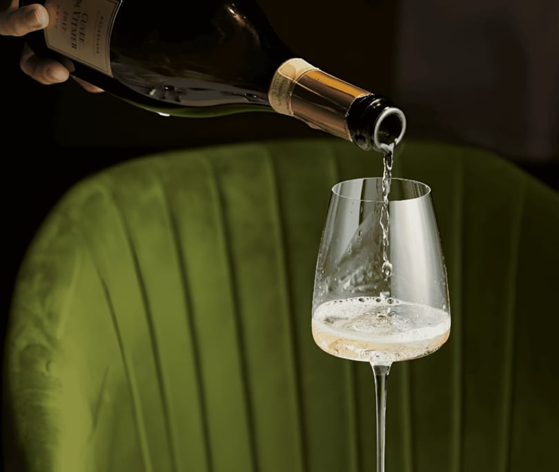 Sparkling wine being poured from a bottle into a glass, with a green velvet chair in the background.