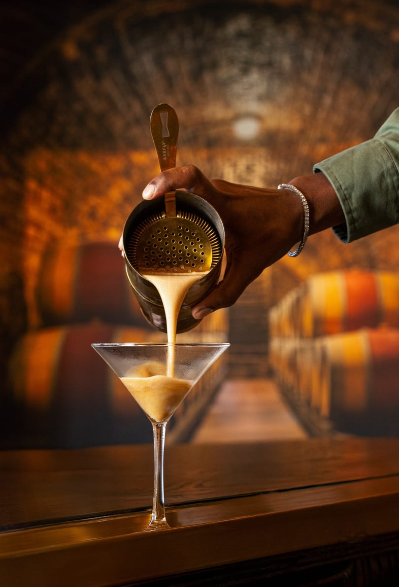 Bartender pouring a creamy cocktail from a shaker into a martini glass on a wooden bar.