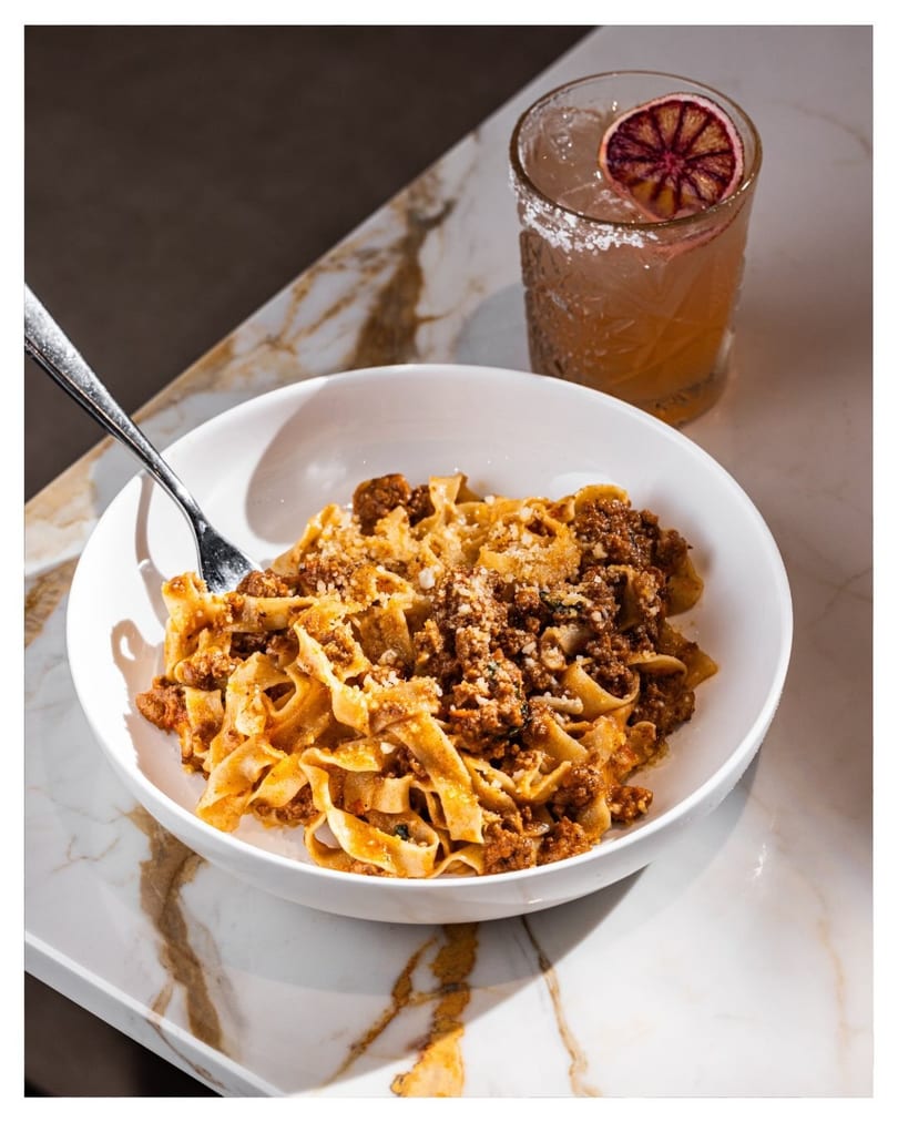 Fettuccine bolognese with parmesan cheese and a cocktail with a blood orange slice on a marble table.